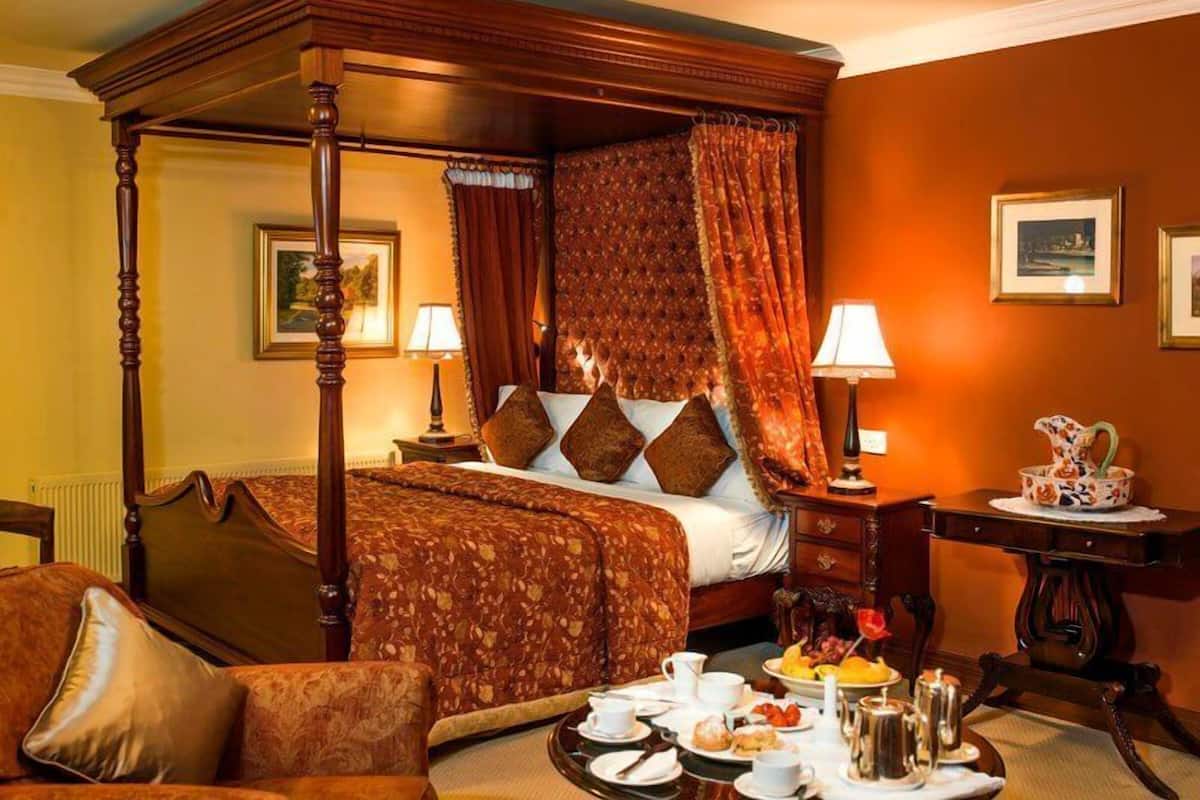 A four-poster bed with a floral bedspread, two bedside lamps, and a dining table set with breakfast.