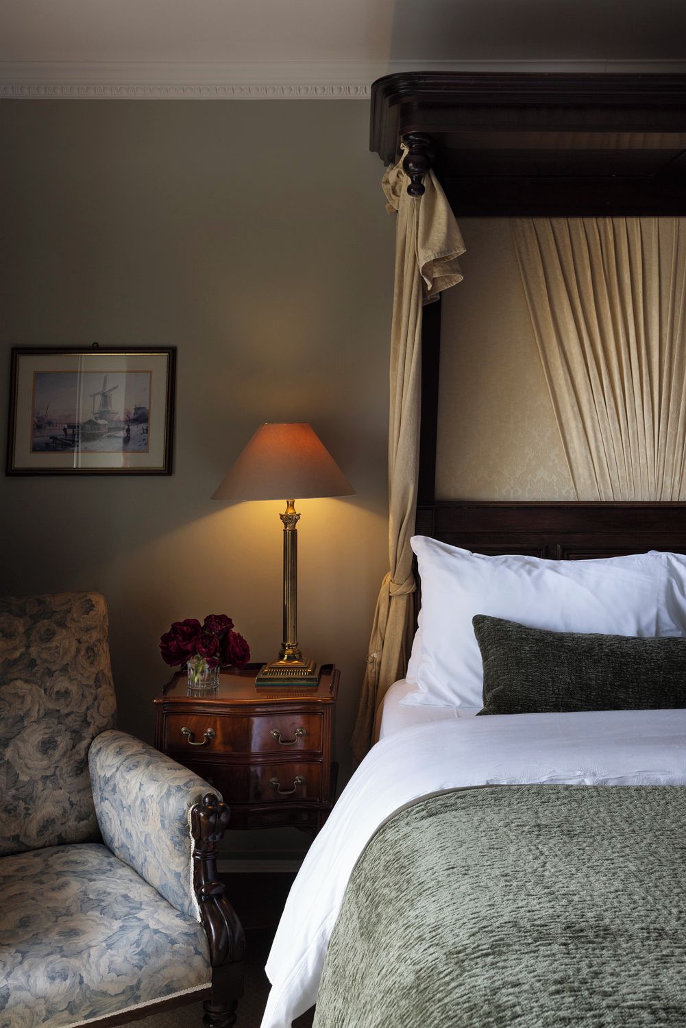 A bedroom with a four-poster bed, a nightstand with a lamp, a chair, and a framed picture on the wall.