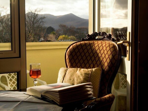 A room with a view of mountains through a window, a chair with a patterned cushion, a glass of red drink, and an open book on a table. - Earls Court House Hotel (Killarney)