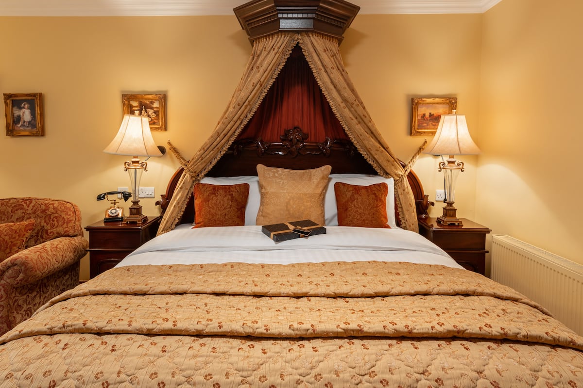 A four-poster bed with a canopy, a classic wooden headboard, and a floral-patterned bedspread. There are two bedside tables with lamps, a chair, and framed pictures on the walls.