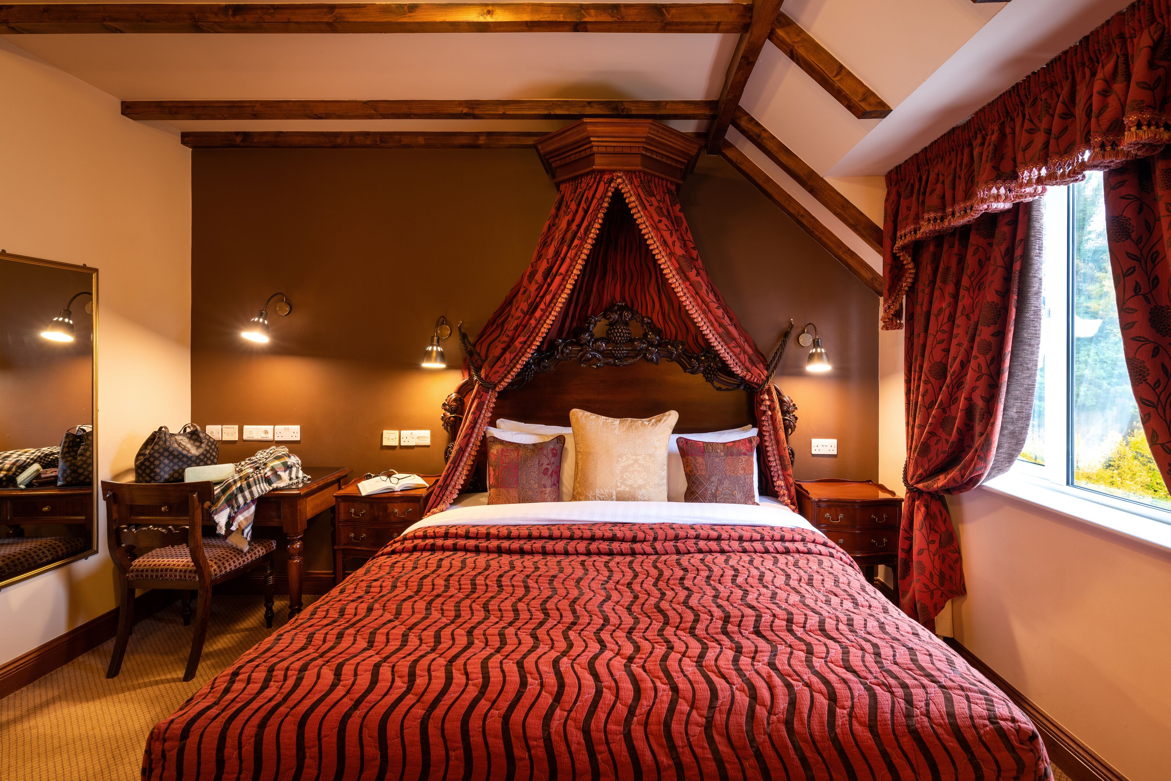 A four-poster bed with a red and black striped bedspread, a wooden headboard, and a canopy. There is a desk with a chair and a lamp, a window with red curtains, and a mirror on the wall.