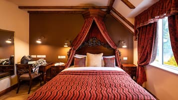 A four-poster bed with a red and black striped bedspread, a wooden headboard, and a canopy. There is a desk with a chair and a lamp, a window with red curtains, and a mirror on the wall.