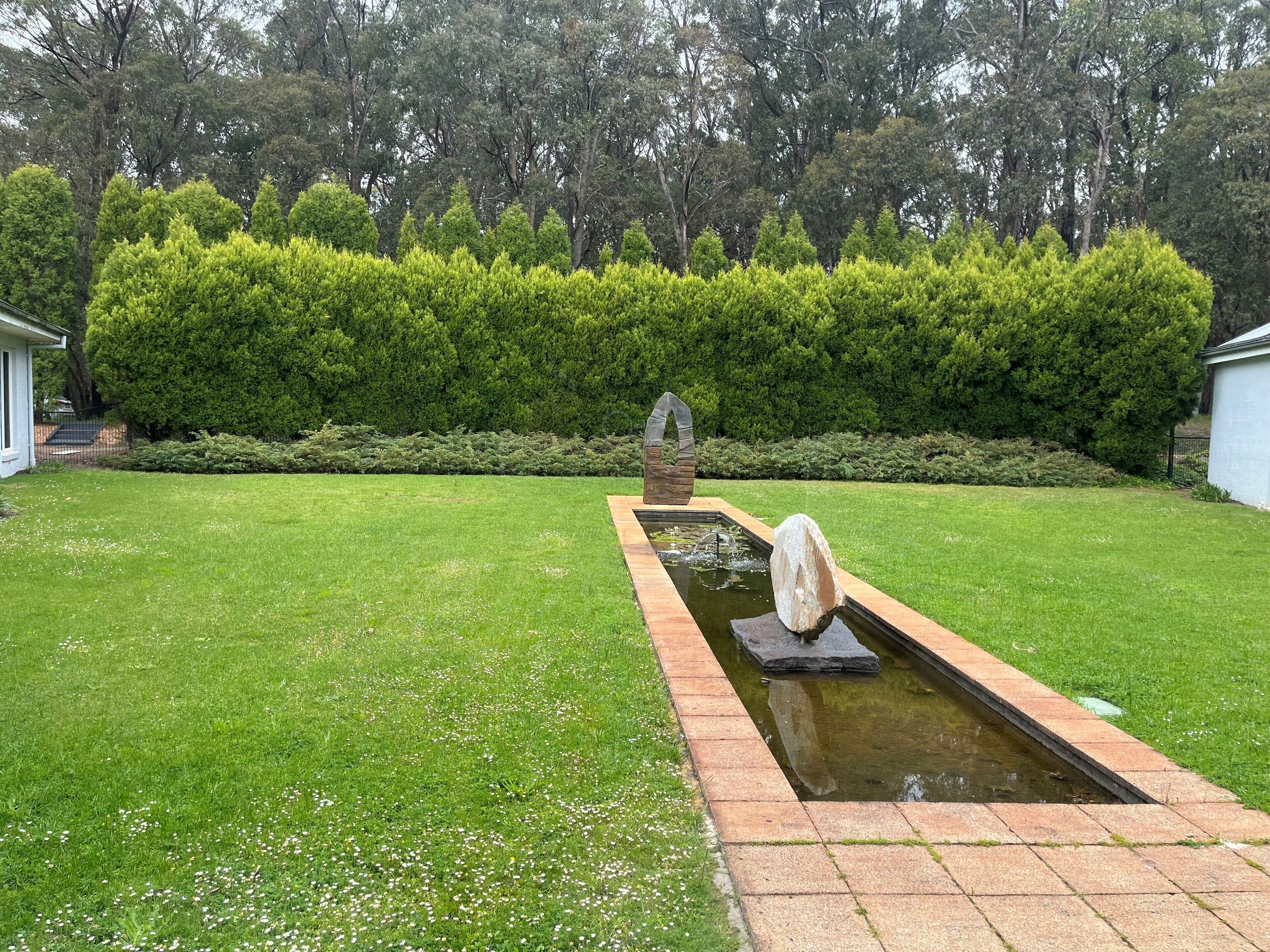 A garden with a rectangular water feature, stone sculptures, and neatly trimmed hedges.