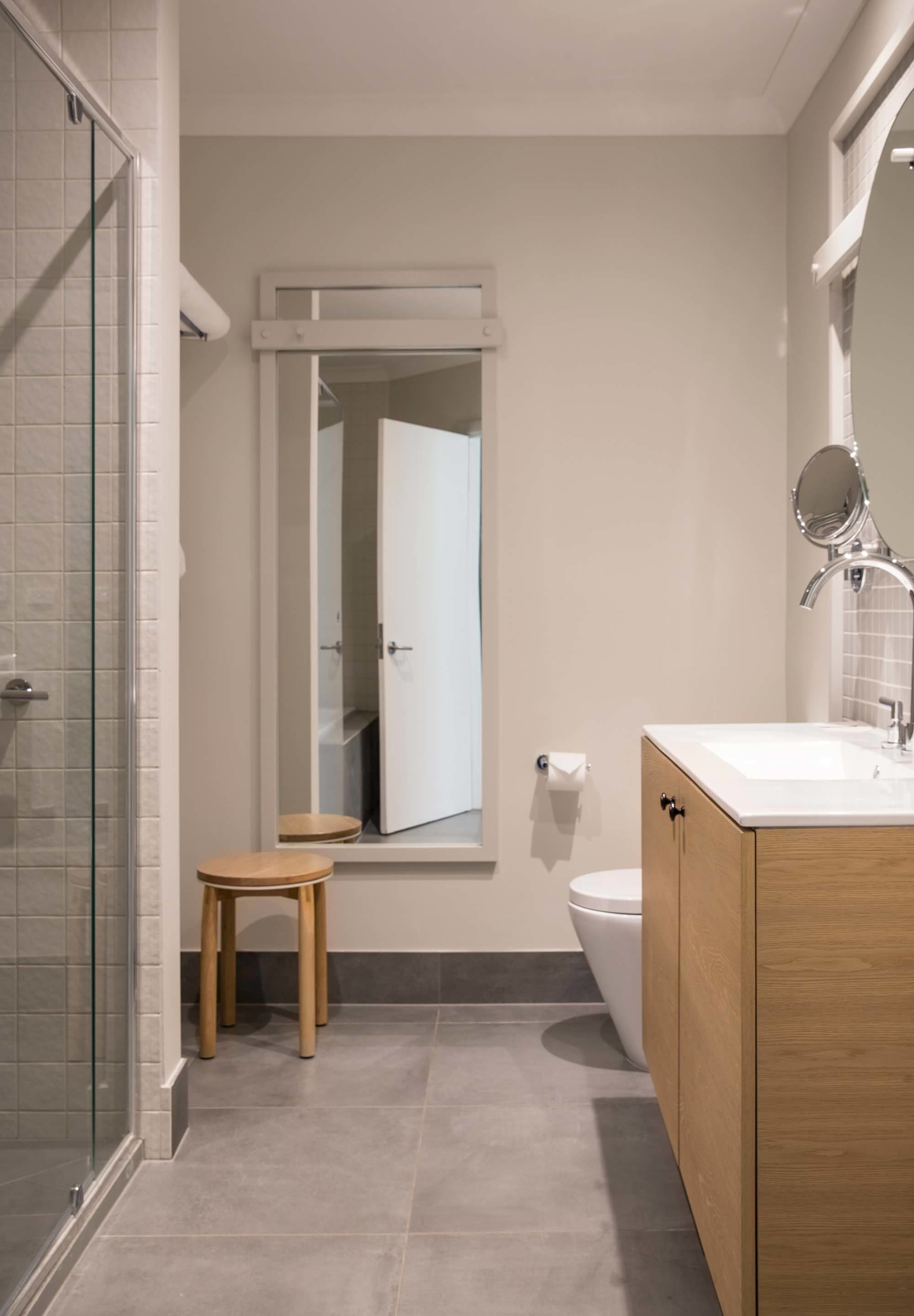 Un bany modern amb un espai de banyera de vidre, un tamboret de fusta, un lavabo blanc amb mirall i un vàter.
