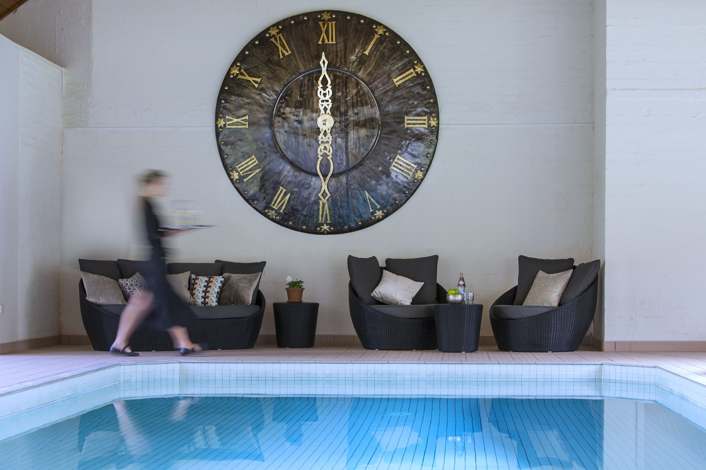 Ein moderner Poolbereich im Innenbereich mit einer großen Wanduhr, bequemen Sitzgelegenheiten und einem gefliesten Poolboden.