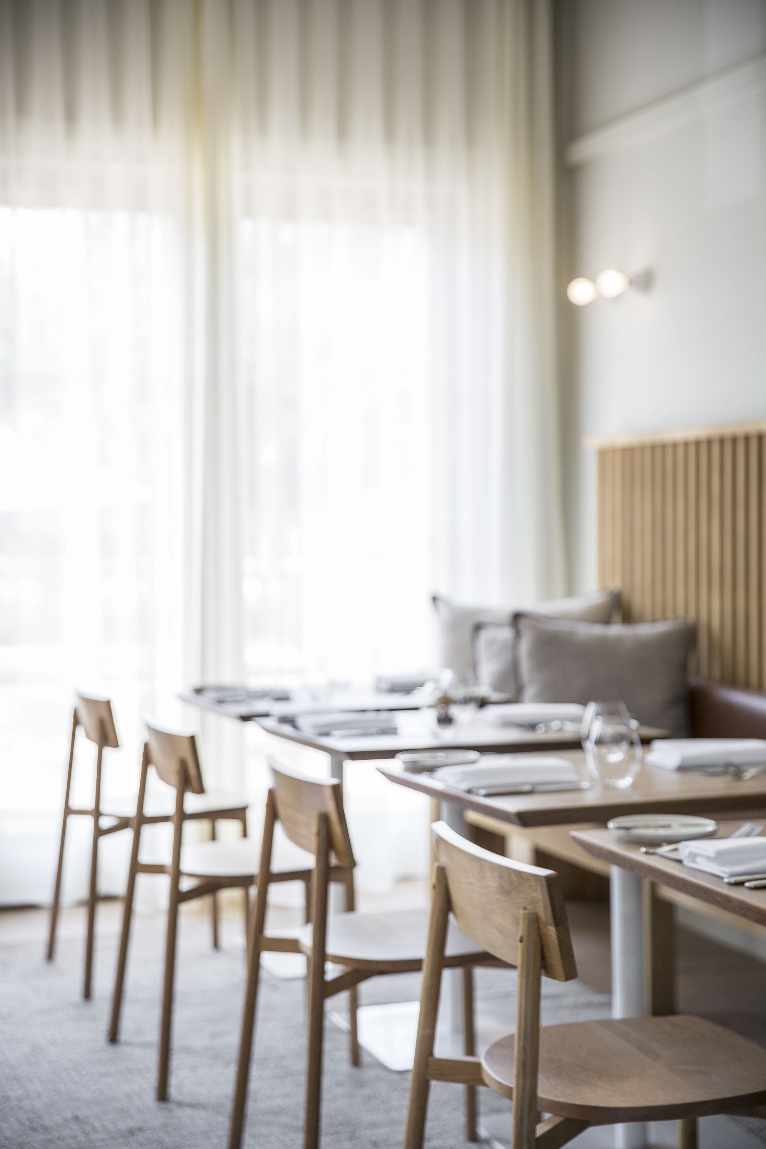 Un comedor moderno con sillas y mesas de madera, ventanas amplias con cortinas ligeras y una paleta de colores neutros.