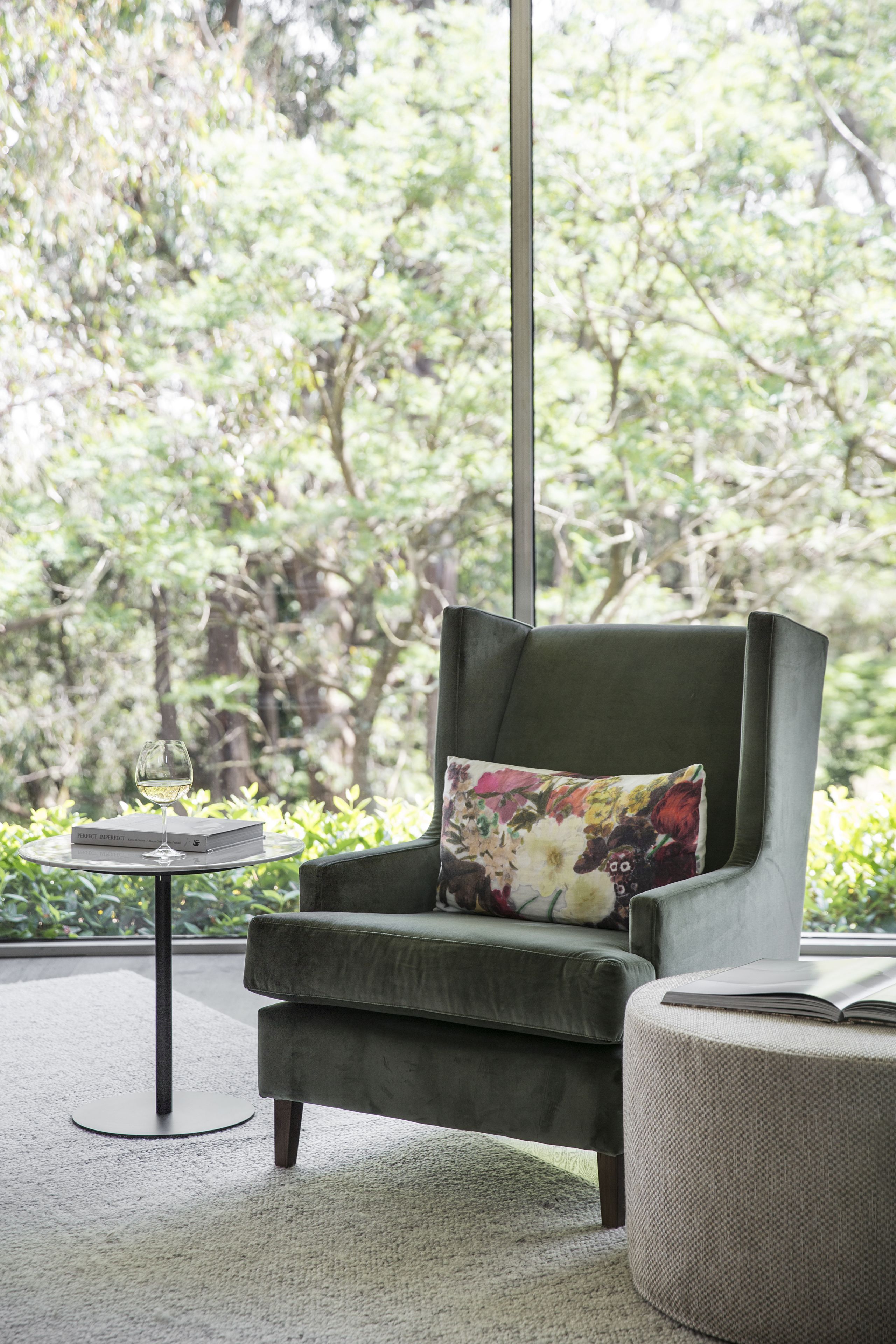 A modern living room with a green armchair, a round ottoman, and a small table with a glass of wine and a book.