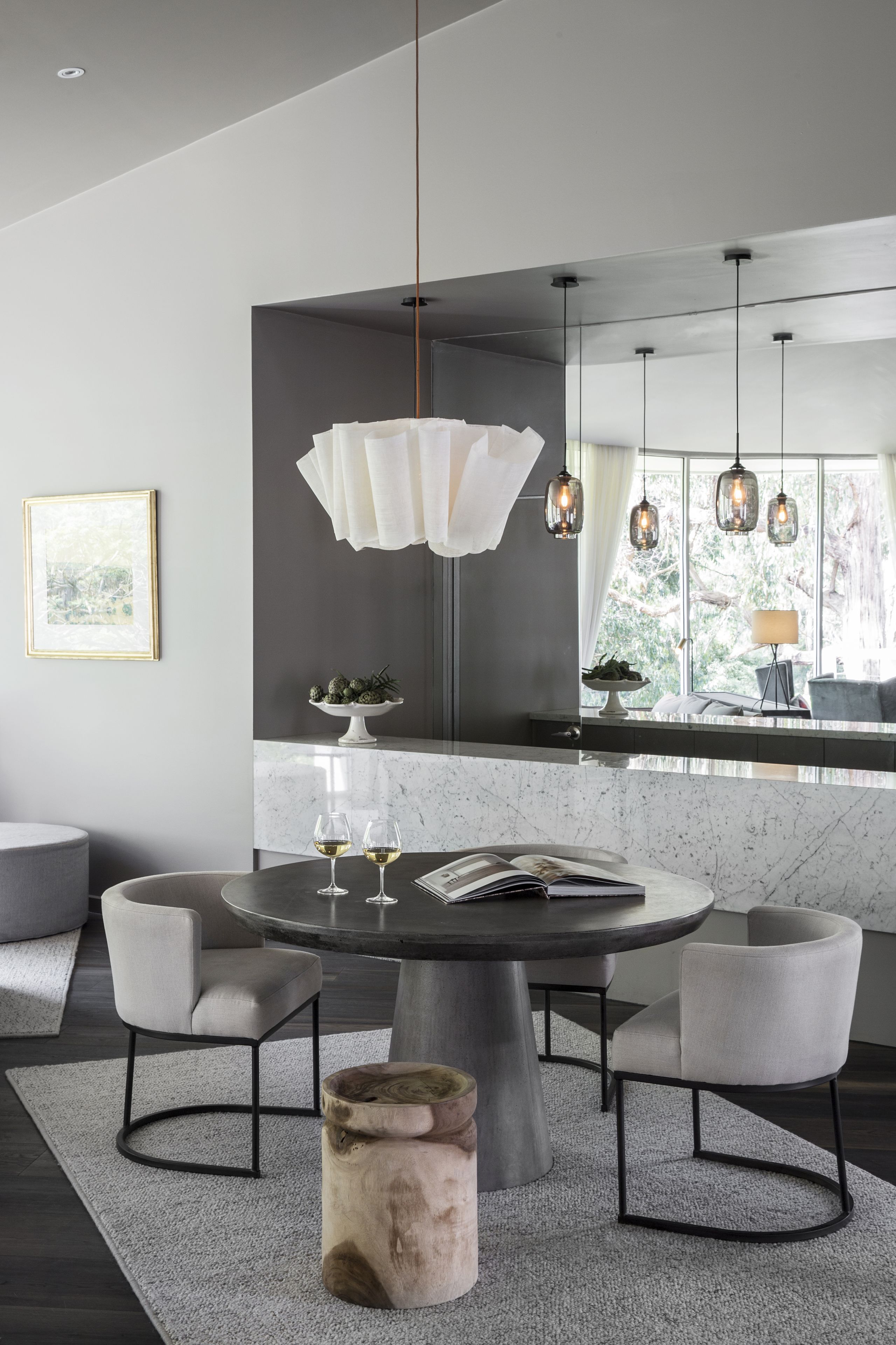 Un espace repas moderne avec une table ronde, quatre chaises, un luminaire central et une grande fenêtre donnant sur un paysage verdoyant.