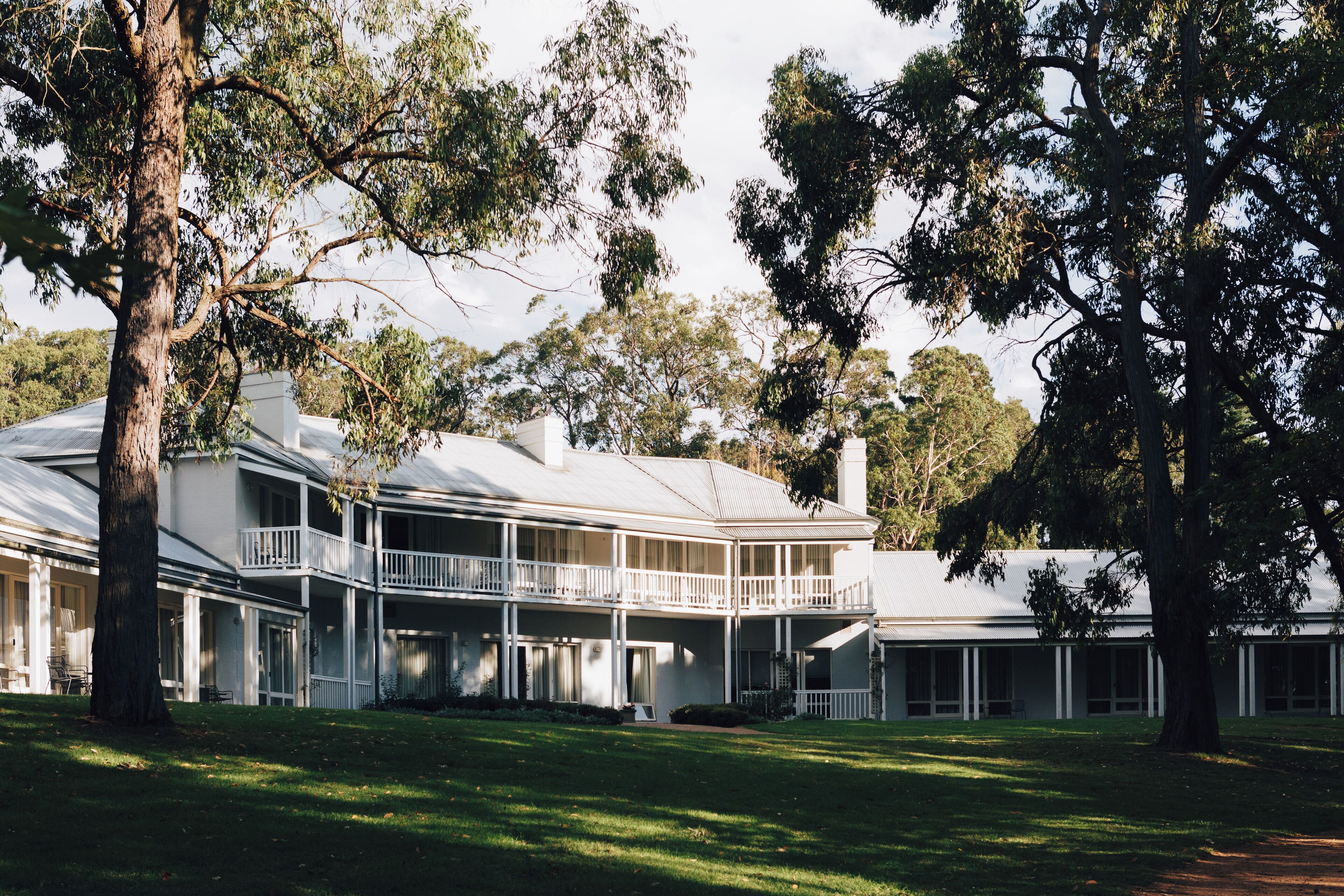A large, two-story building with a balcony on the upper floor, surrounded by trees and a grassy lawn.