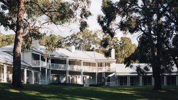 A large, two-story building with a balcony on the upper floor, surrounded by trees and a grassy lawn.