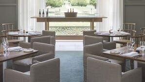 Un comedor con mesas dispuestas para cuatro, un amplio ventanal arqueado con cortinas y vistas a un jardín cuidado.