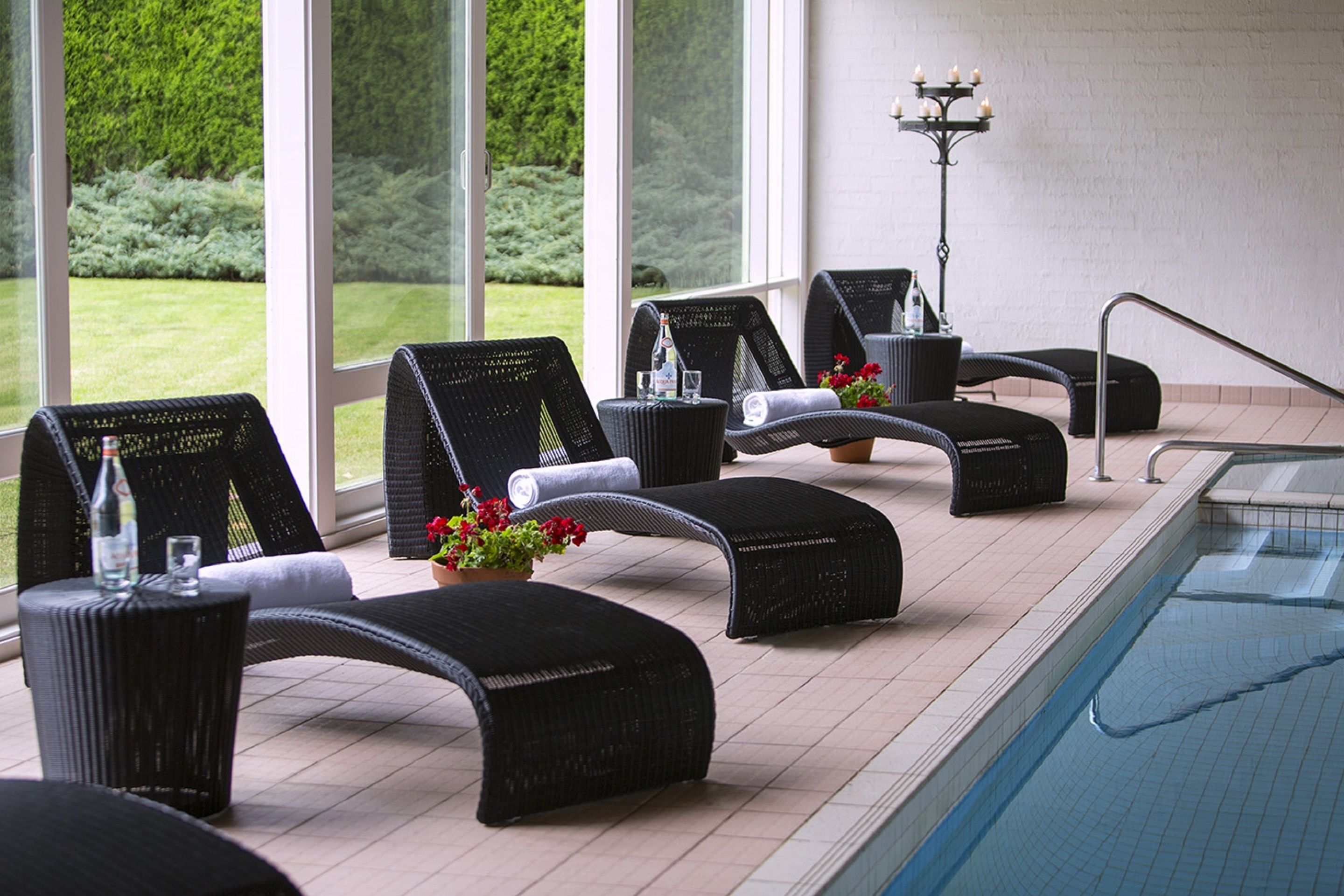 A poolside lounge area with wicker lounge chairs, a small table with a bottle and glass, and a potted plant with red flowers.