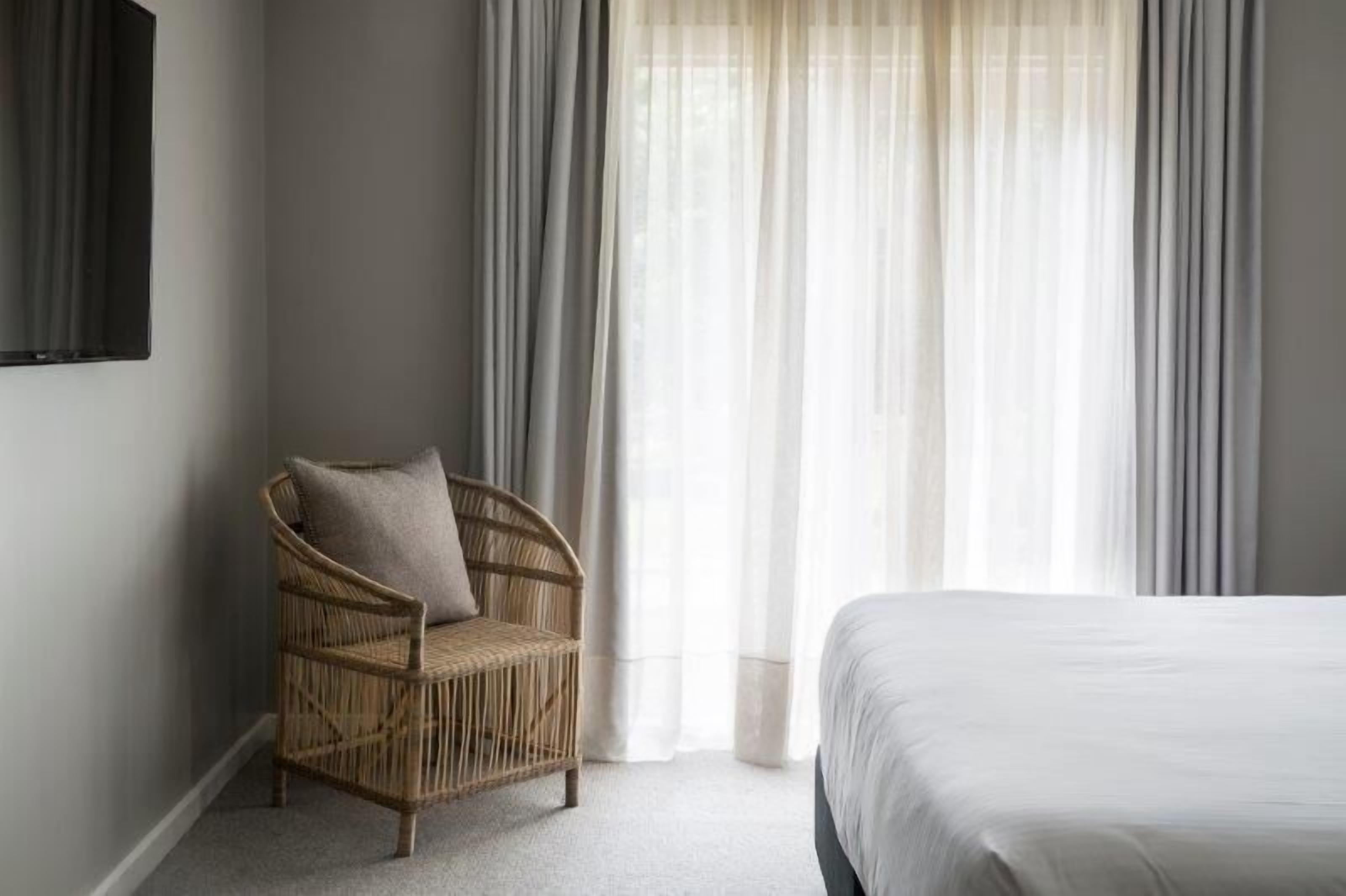 A bedroom with a bed, a wicker chair with a cushion, and sheer curtains.