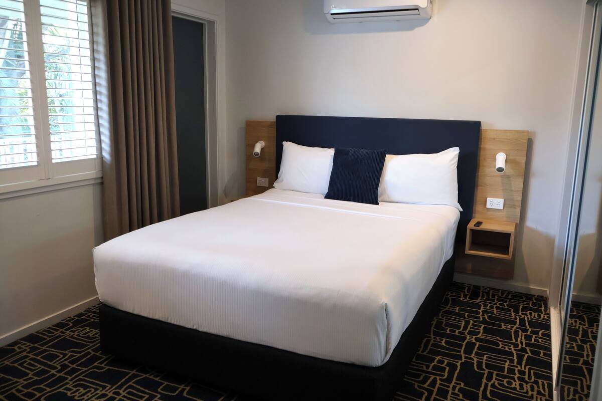Una habitación de hotel con una cama, dos mesitas de noche, un equipo de aire acondicionado y una ventana con cortinas.