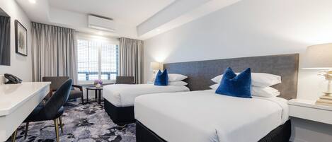 A hotel room with two beds, a desk, a chair, a small table with a vase, and a window with curtains.