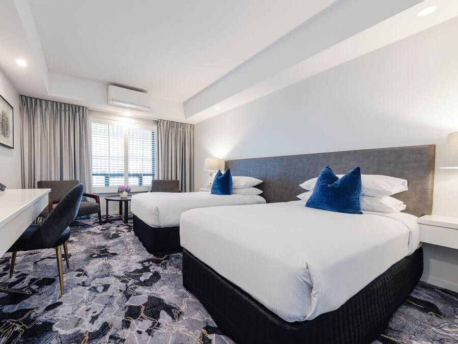 Habitación de hotel con dos camas, un escritorio, una silla, una mesita con un florero y una ventana con cortinas.