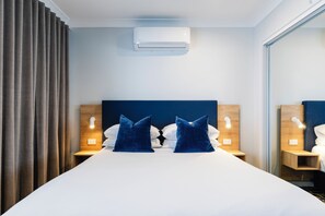 A hotel room with a large bed, two bedside tables, a wall-mounted air conditioner, and a window with curtains. - Hotel Kingsley (Hamilton)
