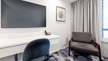 A modern hotel room with a large flat-screen TV mounted on the wall, a desk with a telephone, a comfortable armchair, and a patterned rug on the floor.