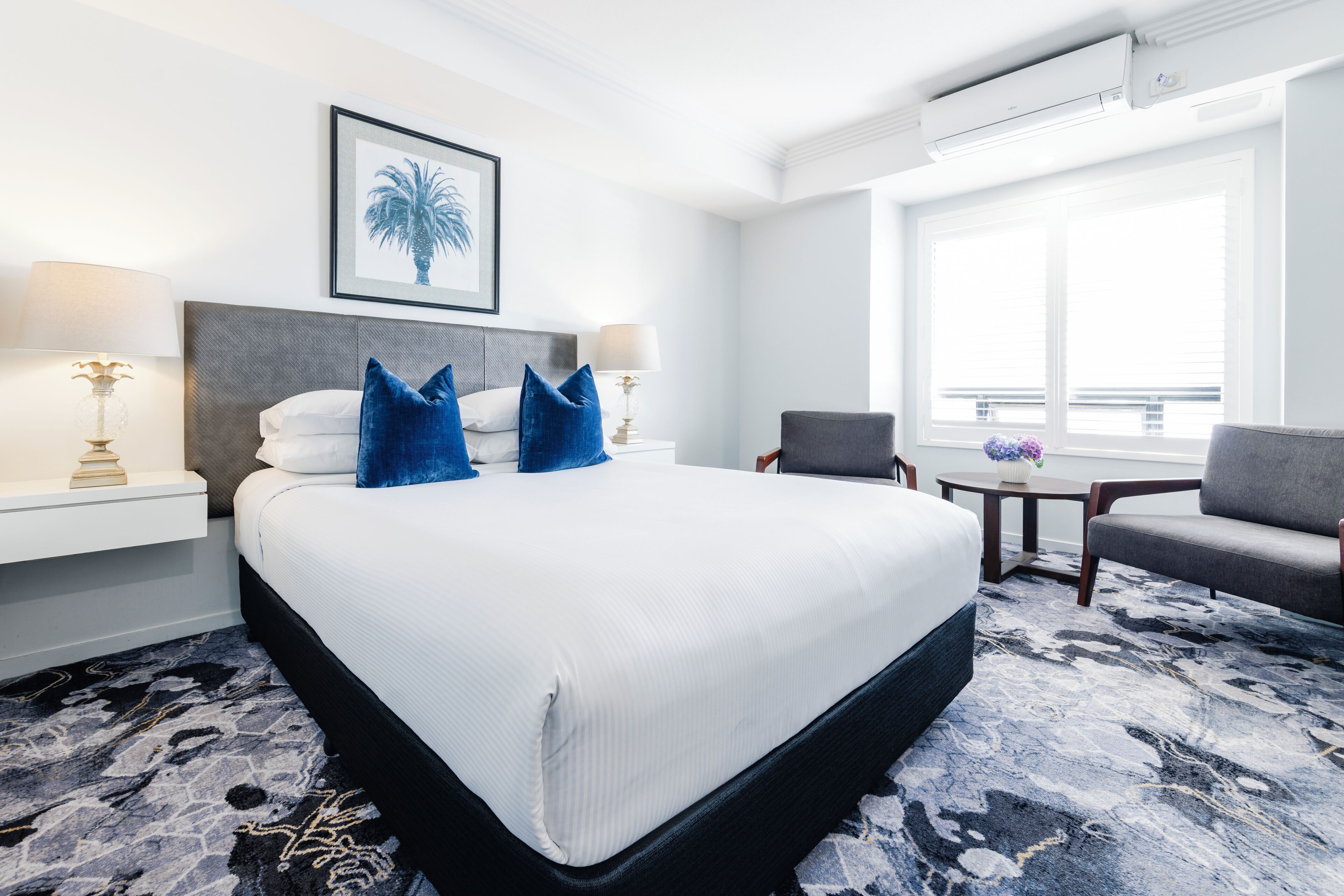 Une chambre d’hôtel avec un grand lit, deux tables de chevet avec des lampes, une chaise, une petite table avec un vase et une fenêtre avec des stores.