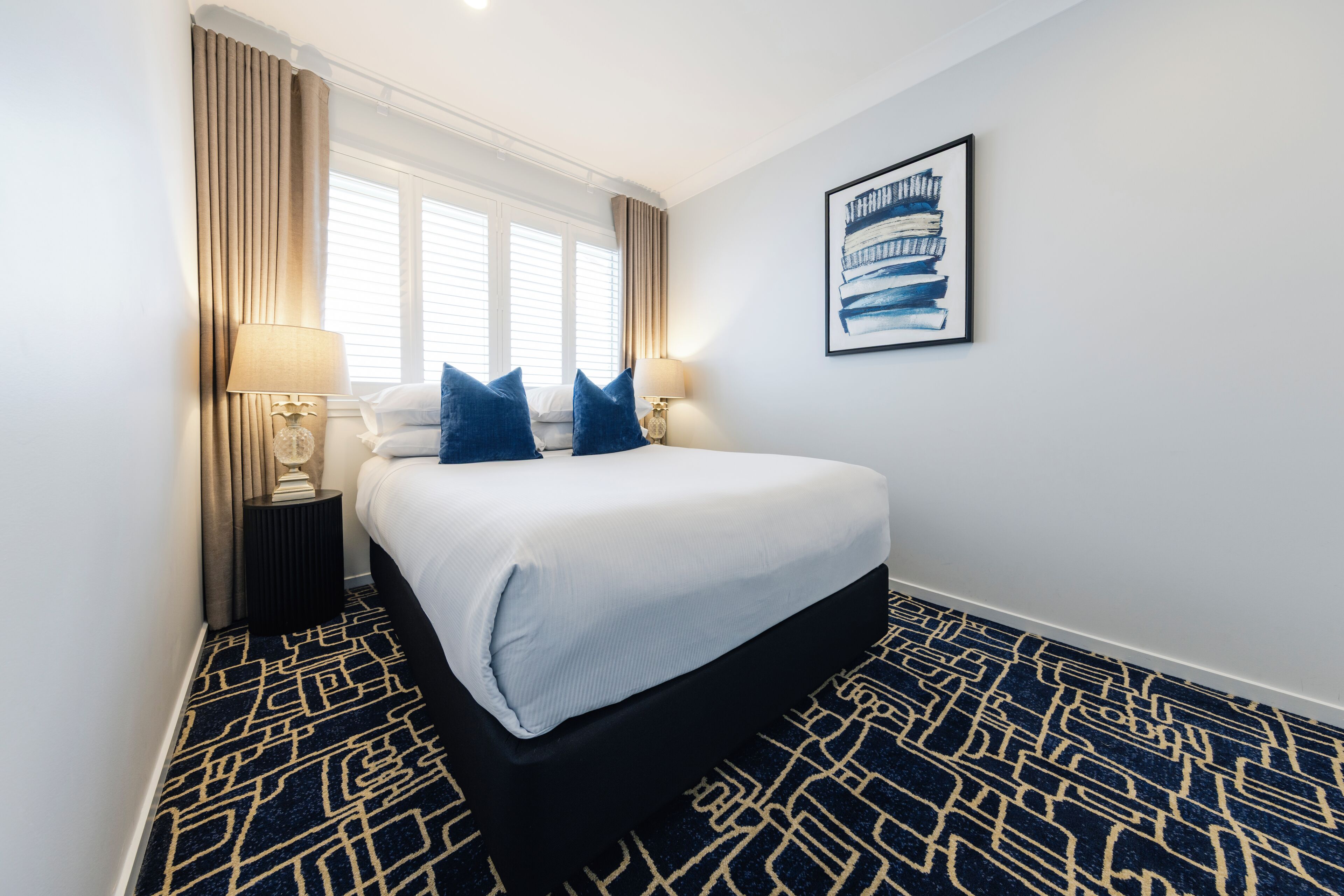 A hotel room with a bed, two bedside lamps, a window with curtains, a wall-mounted artwork, and a patterned carpet.