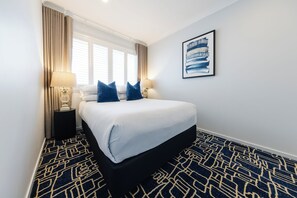 A hotel room with a bed, two bedside lamps, a window with curtains, a wall-mounted artwork, and a patterned carpet. - Hotel Kingsley (Hamilton)