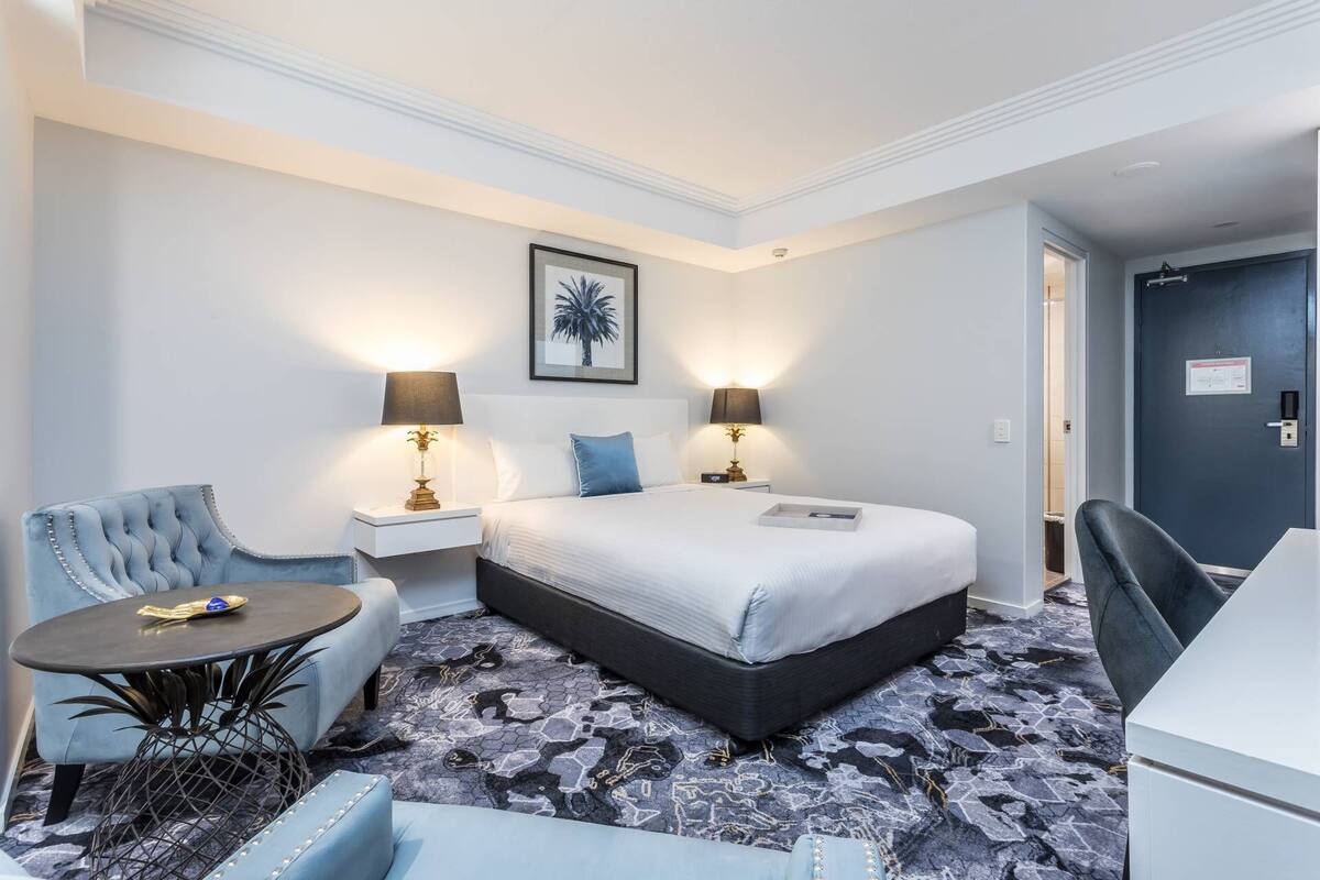 Habitación de hotel con una cama grande, dos mesitas de noche con lámparas, una silla, una pequeña mesa y una alfombra estampada.