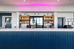 A bar and kitchen area with a neon sign, shelves of alcohol, and a refrigerated display.