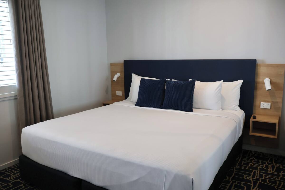 Una habitación de hotel con cama, dos mesitas de noche, una lámpara de pared y una ventana con cortinas.