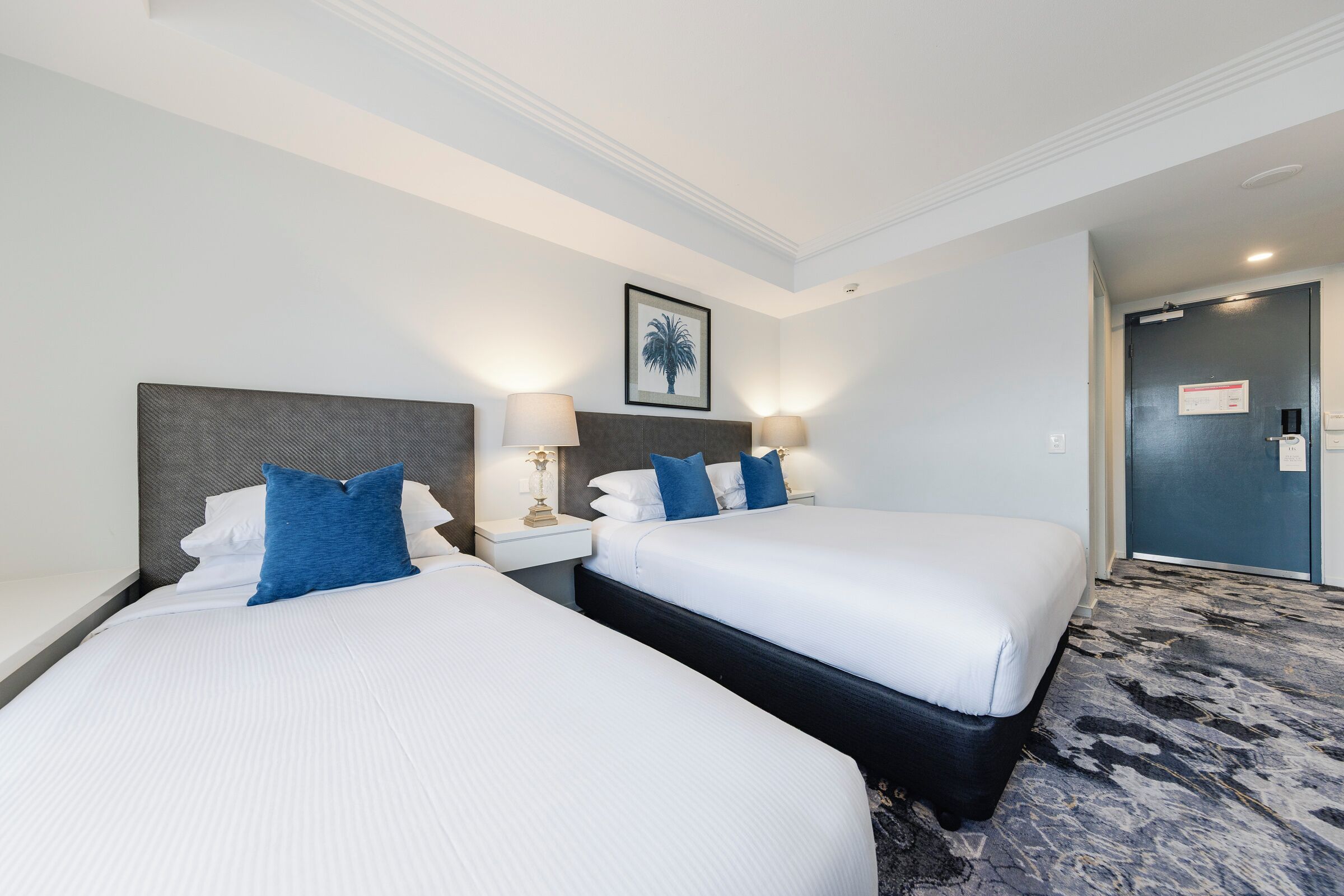 A hotel room with two beds, each with white linens and blue pillows, a nightstand with a lamp, and a framed picture on the wall.