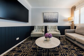 A modern living room with a flat-screen TV, two tufted armchairs, a round coffee table with a vase of flowers, and a patterned carpet. - Hotel Kingsley (Hamilton)