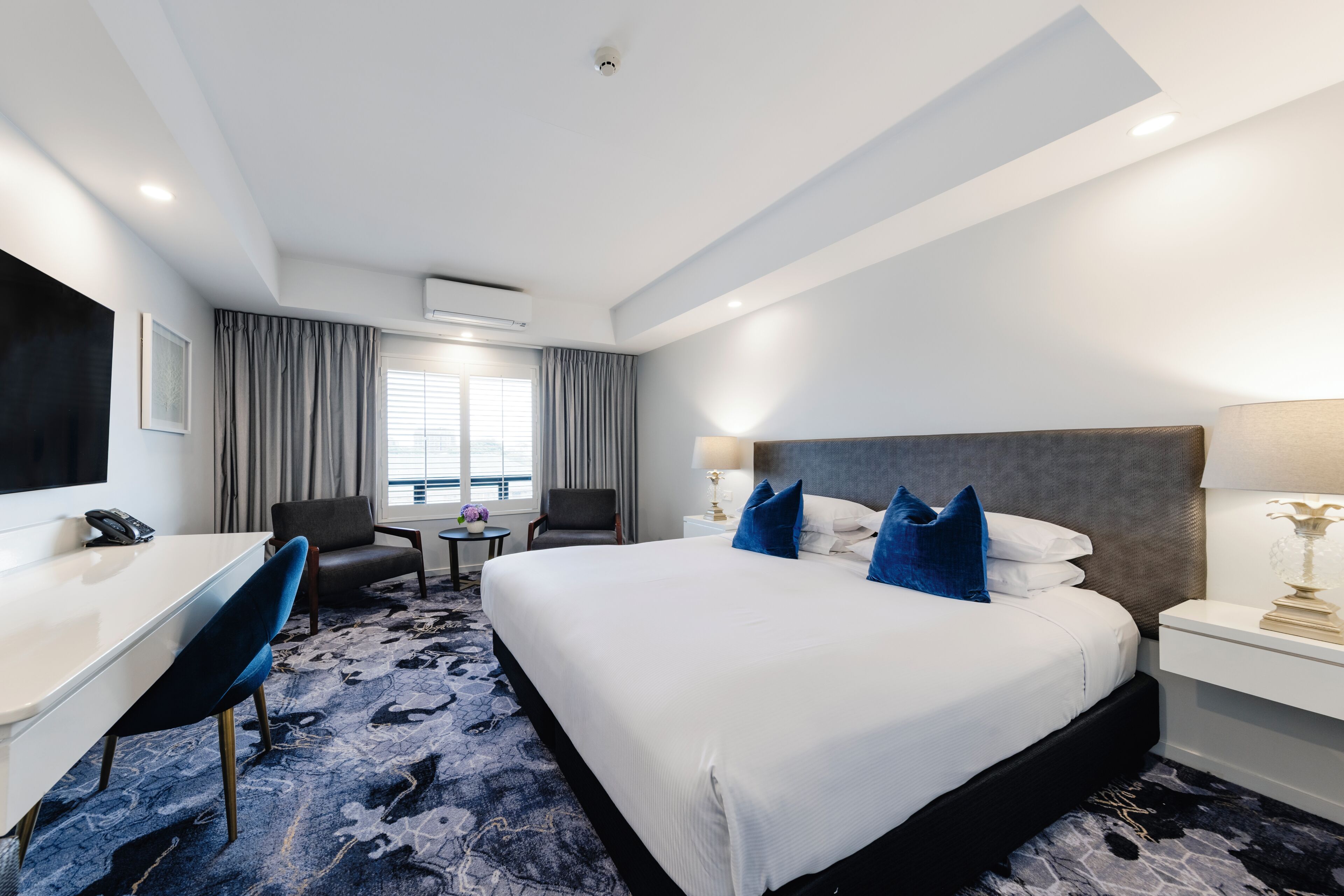Una habitación de hotel con cama, escritorio, silla, televisión y ventana con cortinas.