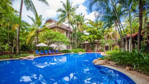 A swimming pool with lounge chairs and umbrellas, surrounded by palm trees and a building with a tiled roof. - Settha Palace Hotel (Vientiane)