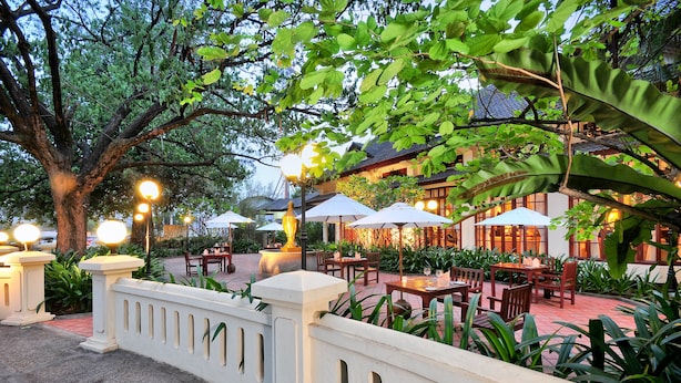 An outdoor dining area with tables and chairs, surrounded by lush greenery and a traditional building in the background. - Settha Palace Hotel (Vientiane)
