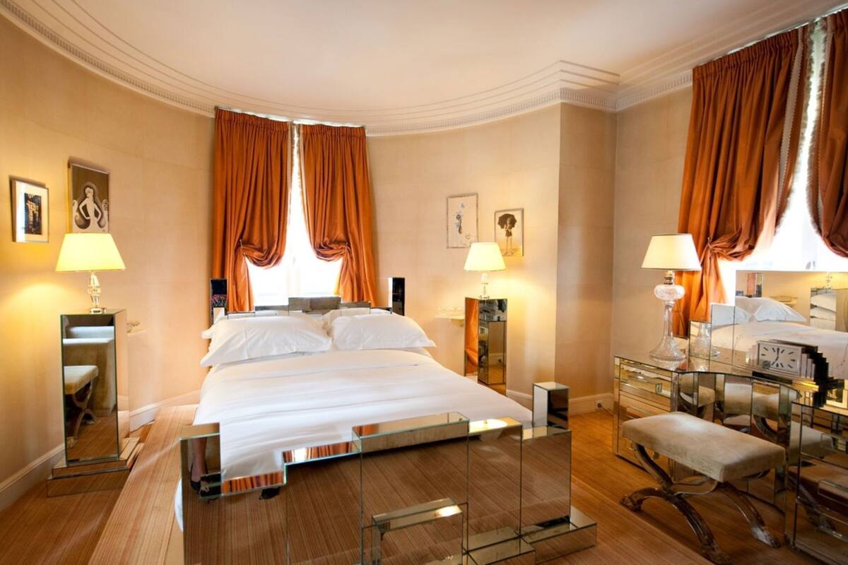 A hotel room with a large bed, two bedside tables with lamps, a mirror, a desk with a chair, and a small bench.