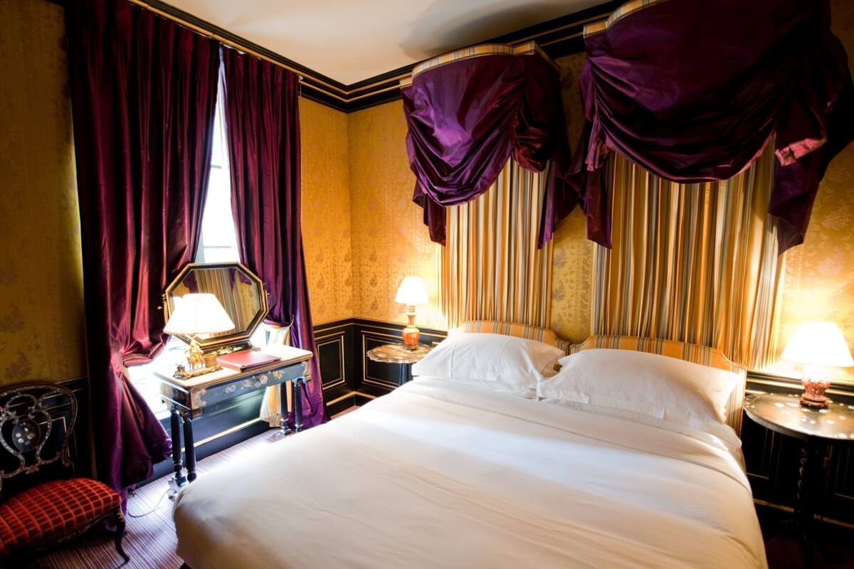 A hotel room with a large bed, two bedside tables with lamps, a mirror, a red chair, and heavy drapery.