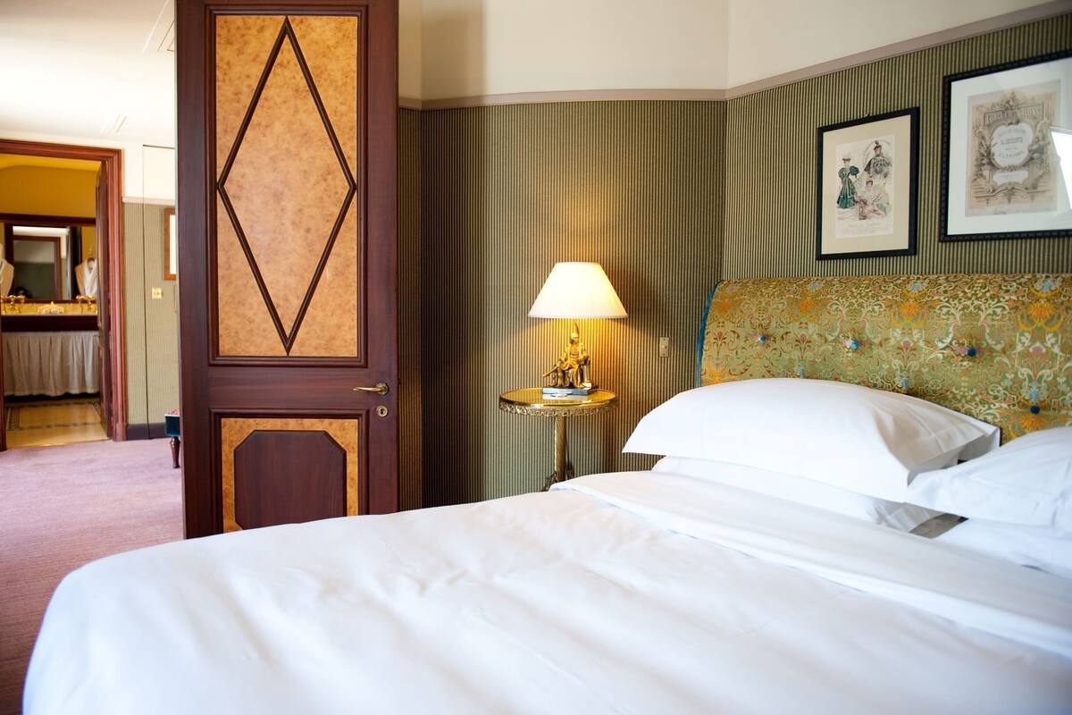 A hotel room with a bed, a nightstand with a lamp, a door with a geometric pattern, and a wall with framed pictures.