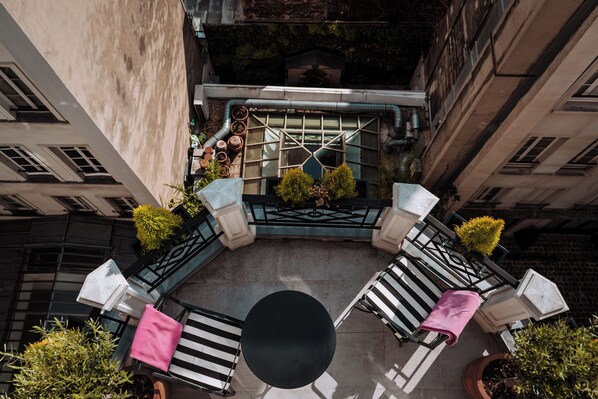 A rooftop terrace with a round table and two lounge chairs, surrounded by potted plants and a decorative metal railing.