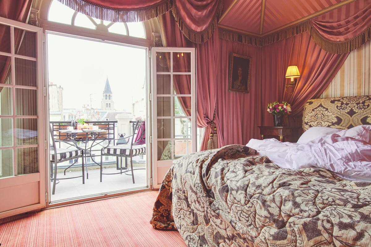 A hotel room with a large bed, a table with chairs, and a view of a cityscape through an open door.