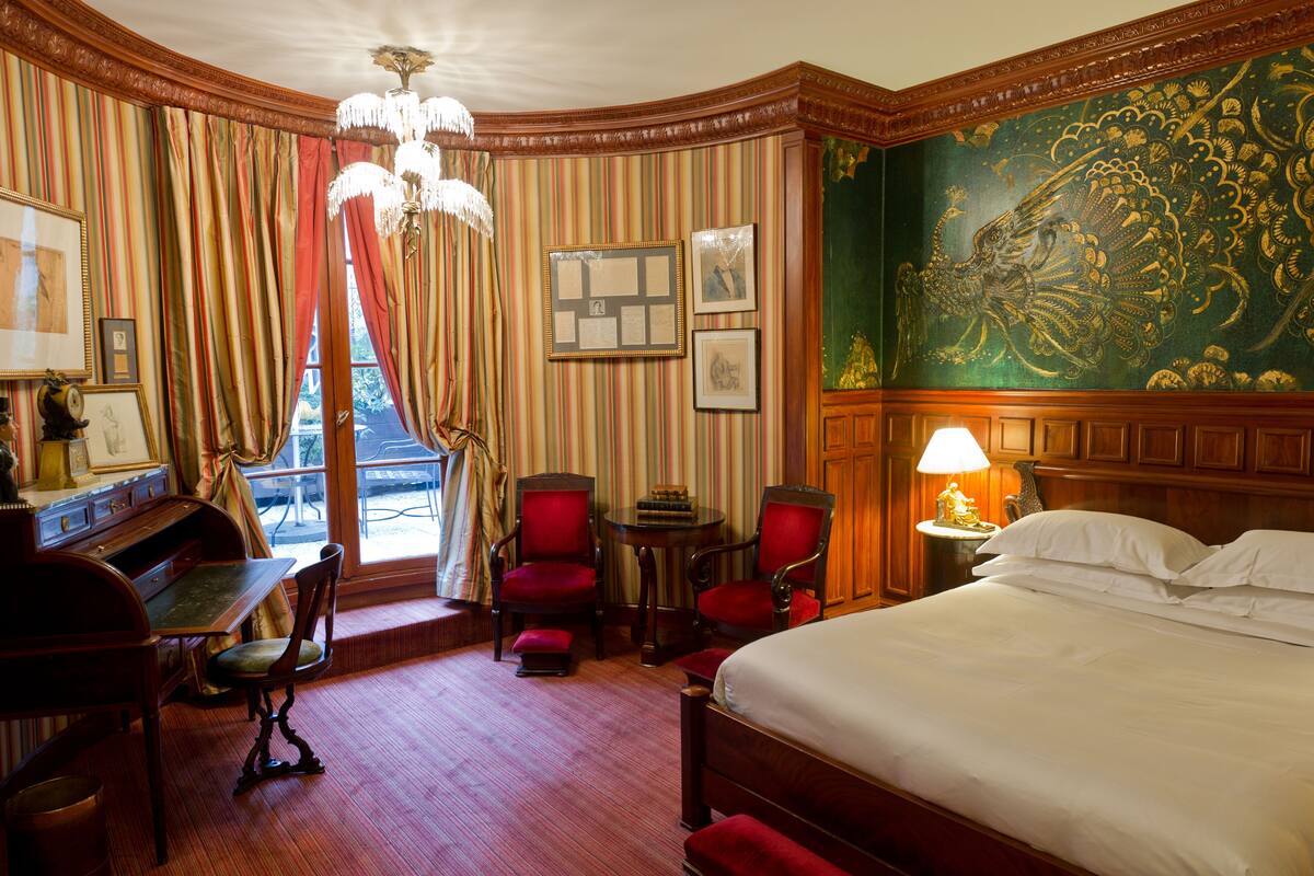 A hotel room with a large bed, a desk with a chair, two armchairs, a small table, and a chandelier.