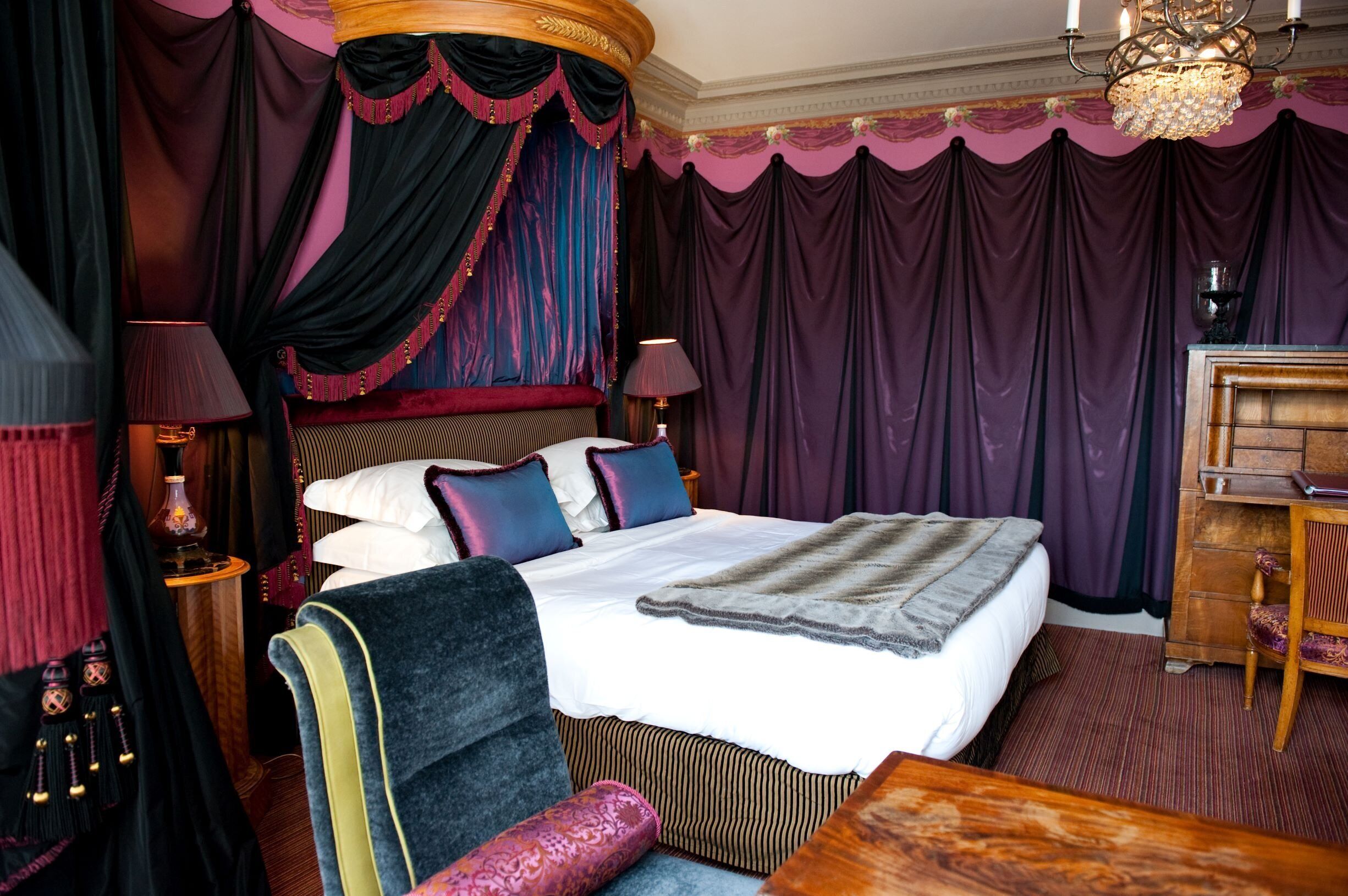 Une chambre à coucher luxueuse, dotée d’un grand lit, de rideaux richement décorés, d’un lustre et d’un bureau en bois.