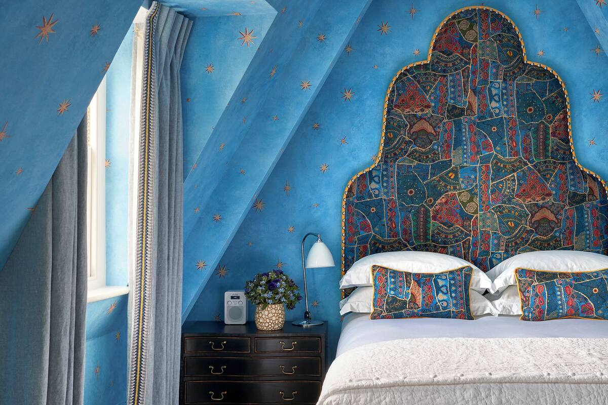 A bedroom with a blue star-patterned wall, a bed with patterned pillows, a nightstand with a radio and a vase of flowers, and a window with curtains.