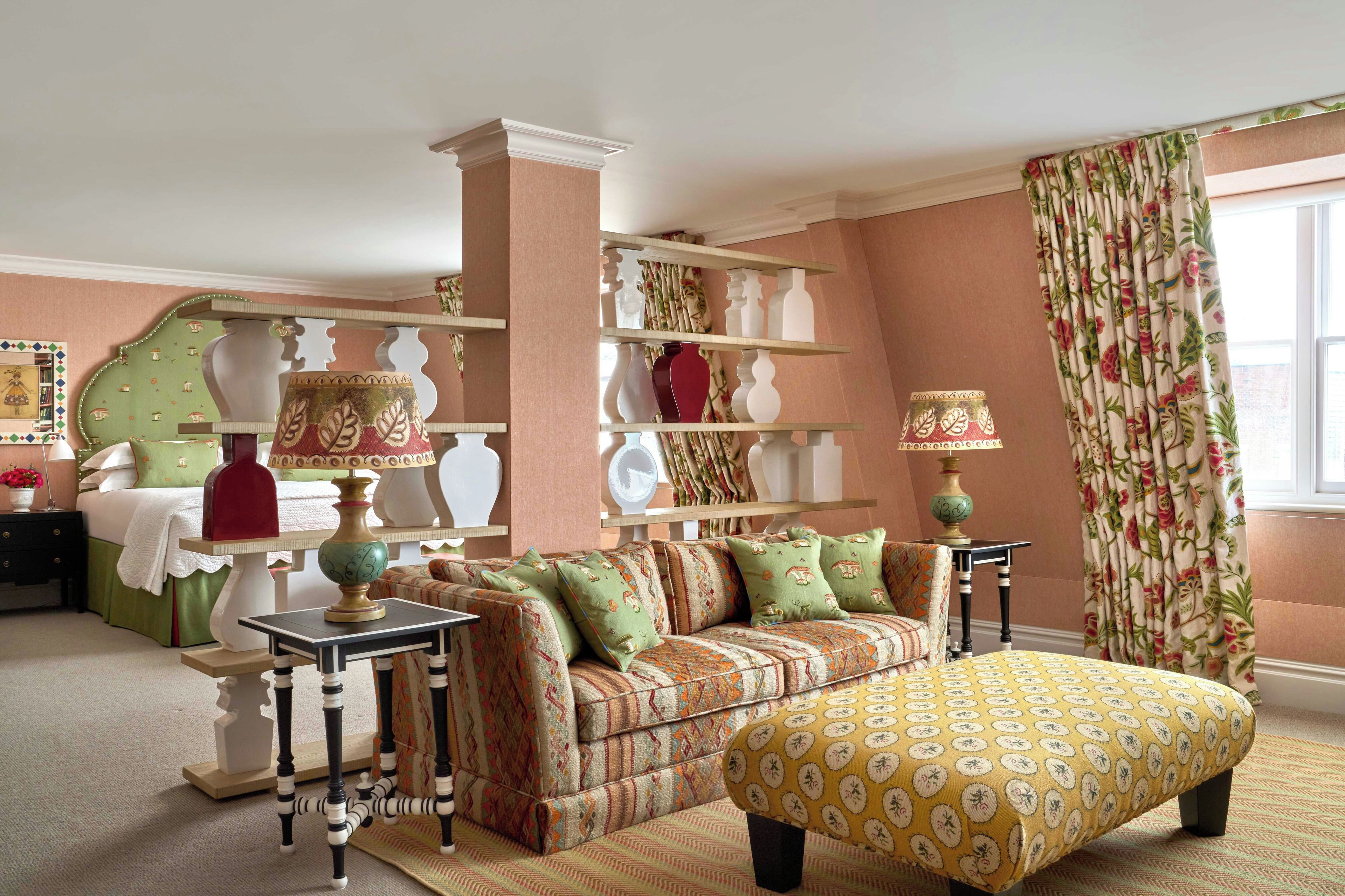 Quarto aconchegante com sofá, banquinho e luminárias. Prateleiras exibem objetos decorativos e uma janela com cortinas.