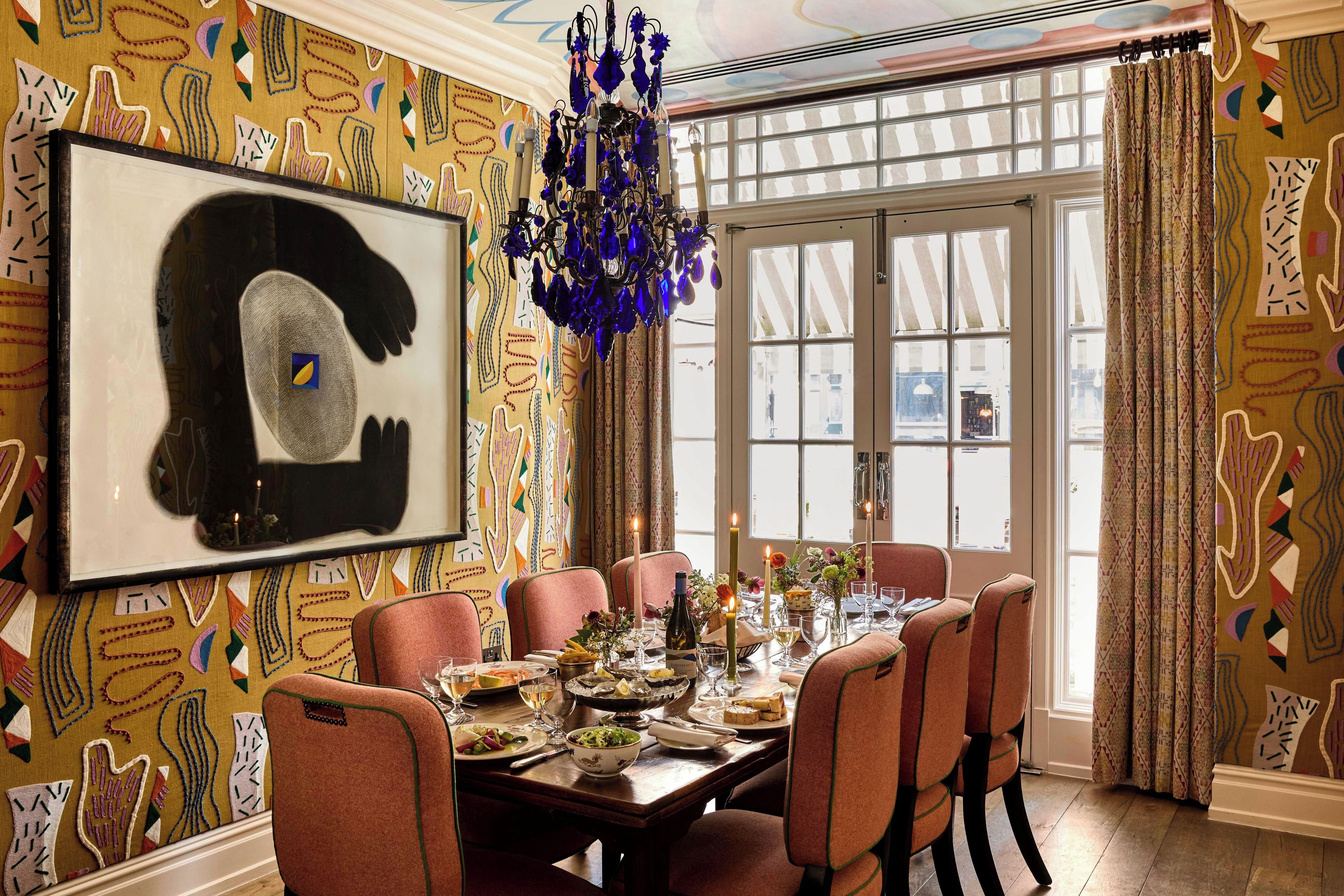 A dining room with a table set for six, a chandelier, and patterned wallpaper.