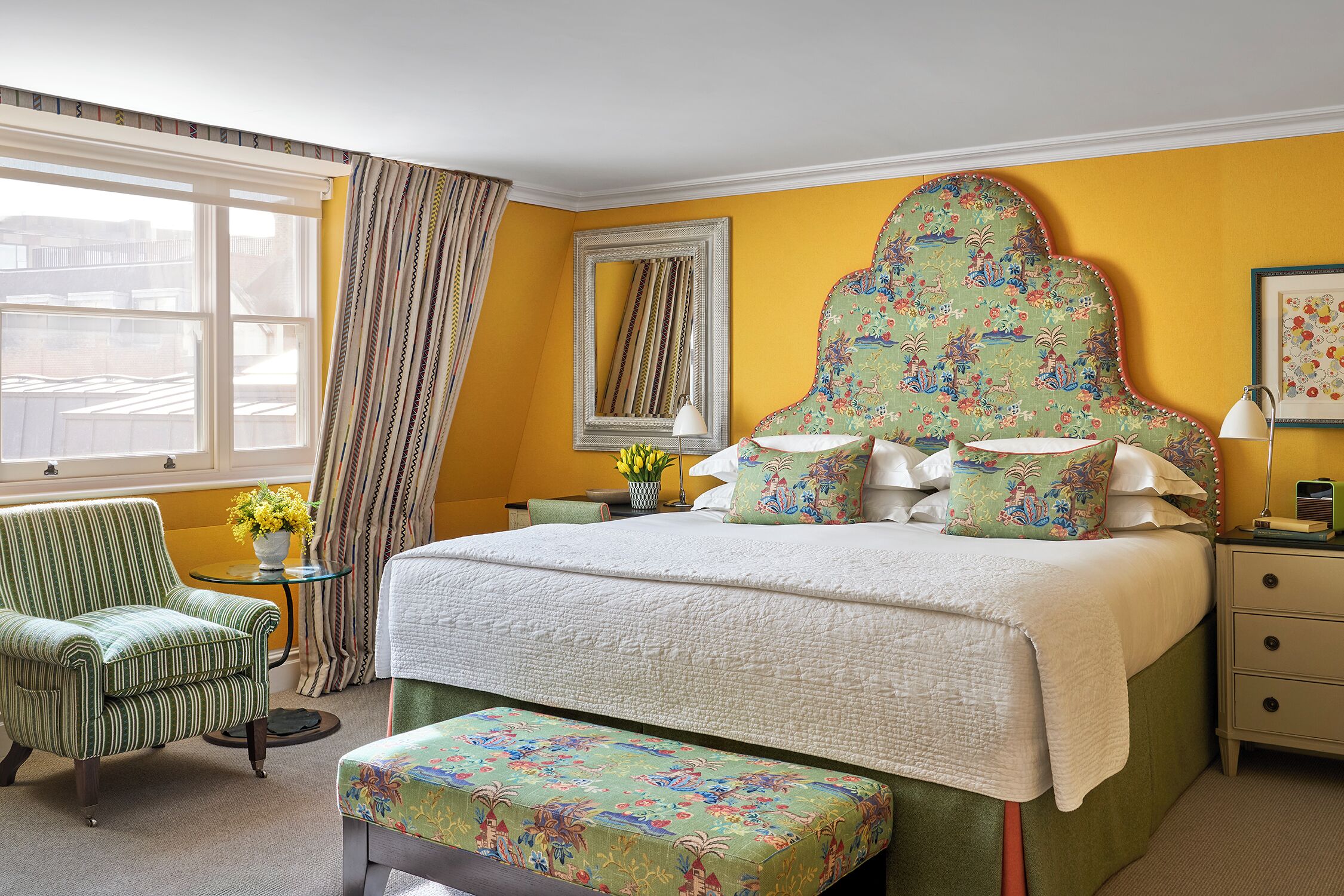 A hotel room with a bed, a green upholstered armchair, a small round table with a vase of flowers, and a window with curtains.