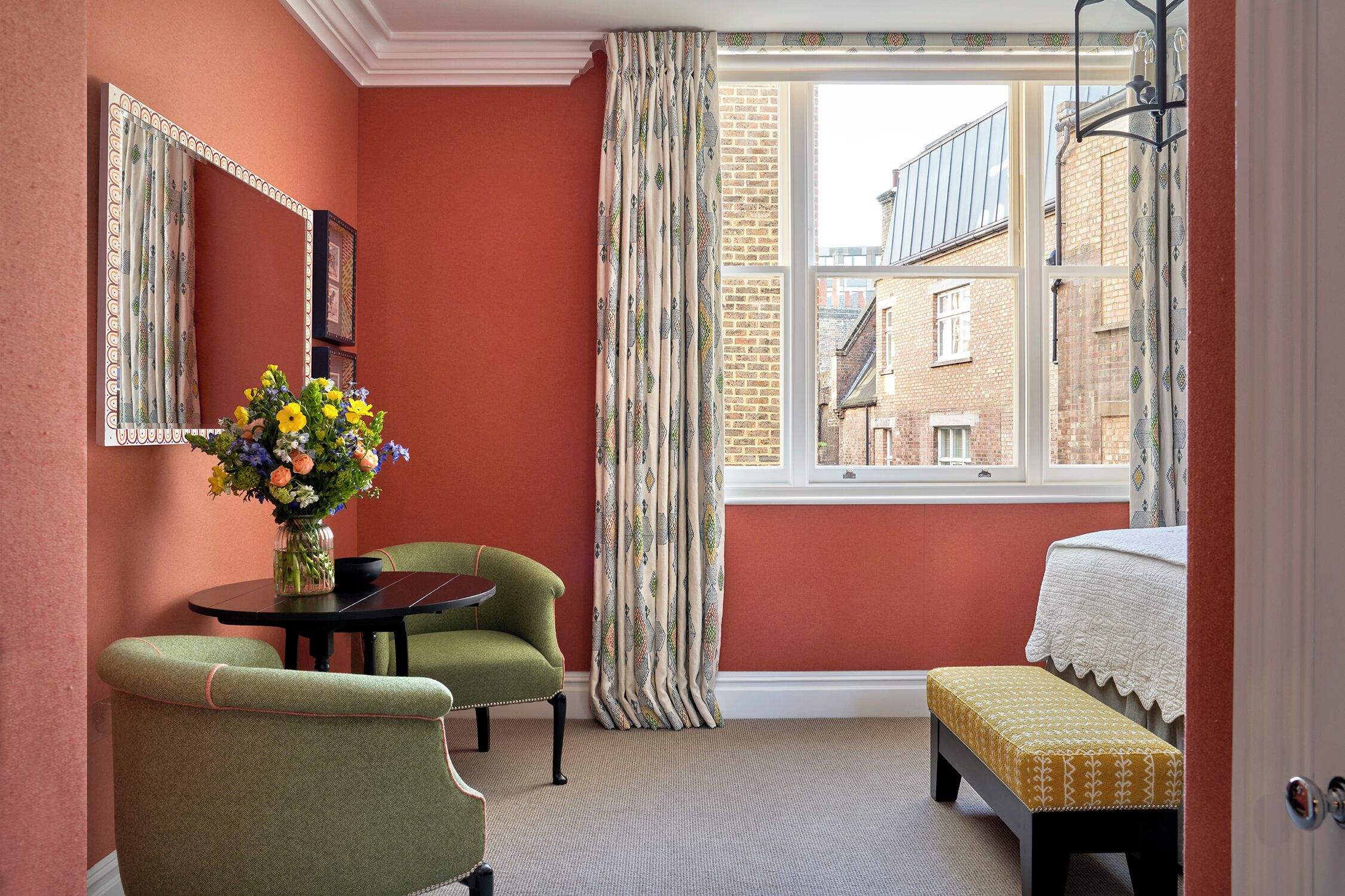 A room with a red wall, a window with curtains, a bed with a white bedspread, a green armchair, a small round table with a vase of flowers, and a bench with a patterned cushion.
