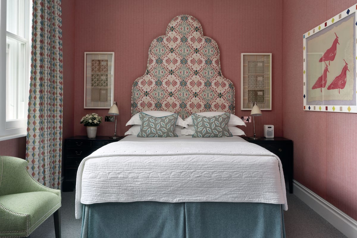 A bedroom with a large bed, a headboard with a floral pattern, two bedside tables with lamps, a green armchair, and a window with patterned curtains.