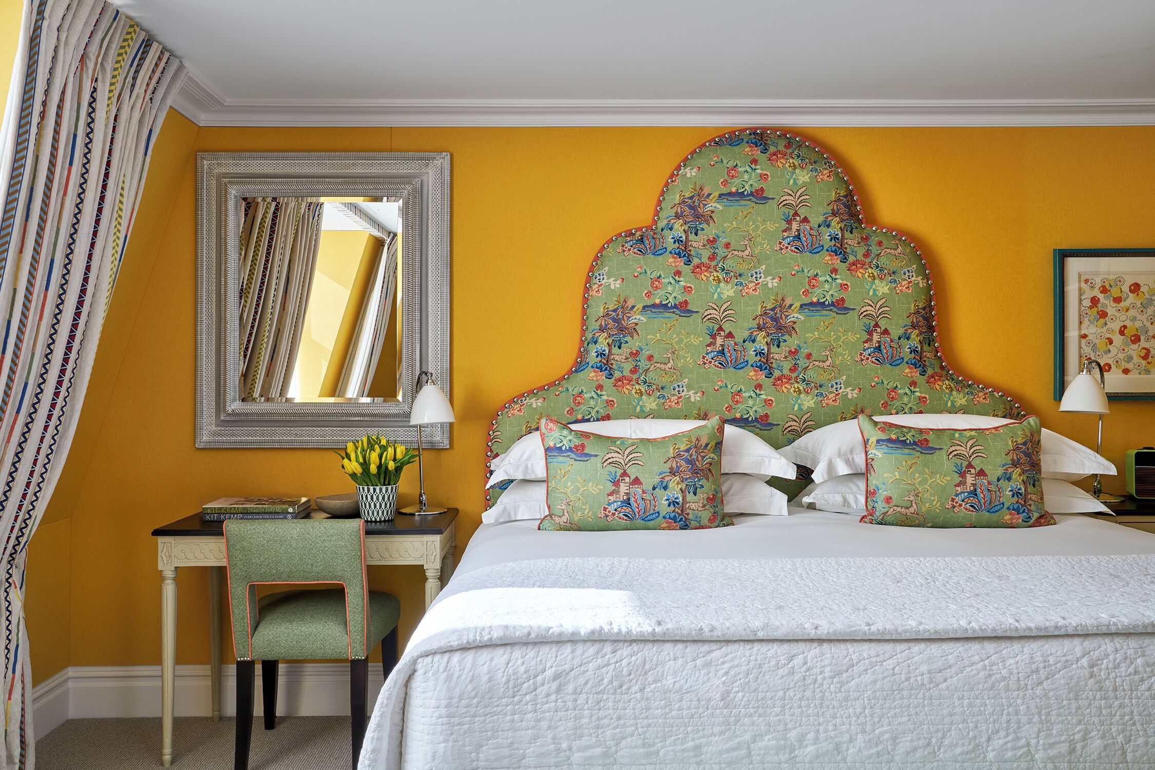 A hotel room with a large bed, a desk with a chair, a mirror, and a floral-patterned headboard.