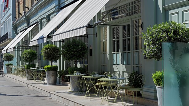 Una fila de mesas y sillas de café al aire libre con macetas, frente a un edificio con toldo blanco y un cartel que dice 'CHILDREN'S CAFE HOTEL'.