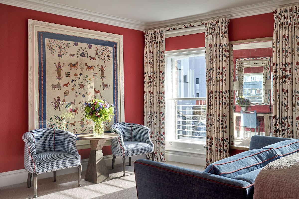 A room with a red wall, a large tapestry, two armchairs, a sofa, and a window with floral curtains.