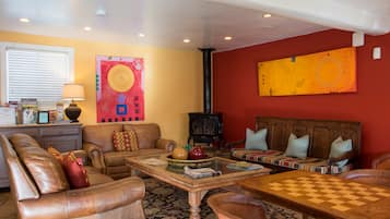 A living room with a sofa, armchairs, a coffee table, and a chess table. There is a fireplace, a window with blinds, and artwork on the walls.